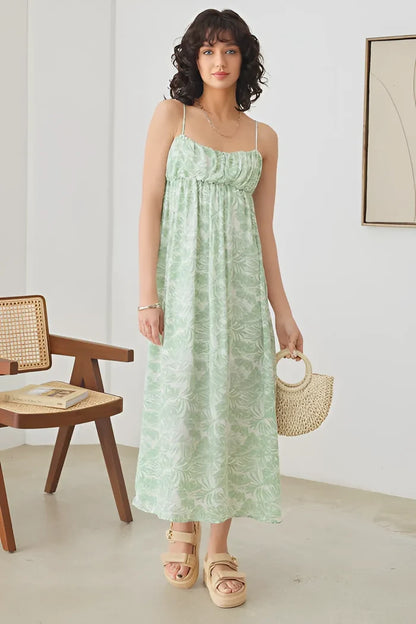 Woman in a green sleeveless shirring tropical print maxi dress standing by a chair